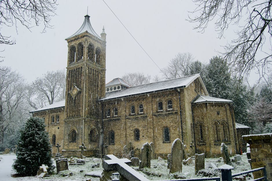 A historic stone church with a tall, square bell tower featuring arched windows and decorative wooden details at the top. The church is situated among snow-covered trees and a graveyard with weathered headstones, some leaning at angles. The snow blankets the roof and the surrounding ground, creating a winter scene. The overcast sky adds a muted, gray tone to the scene, while the church's brick and stone façade displays signs of age and historic architecture. The image captures the quiet atmosphere of a winter day, with no people present, and the church stands as a prominent structure amid the snow-covered landscape, illustrating a typical scene of seasonal change in a residential or historic area, relevant to the context of house removals or property-related services by Walthamstow Man and Van.