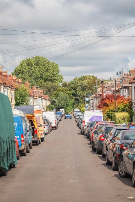 A residential street scene showing a narrow urban road lined with parked cars on both sides, including various sedans, hatchbacks, and vans. The street is situated in a suburban area with terraced houses featuring brick facades, small front gardens, and pitched roofs. Some houses have visible windows and doors, while others are partially obscured by the parked vehicles. In the background, there are lush green trees and a partly cloudy sky with overhead power lines crossing above the street. The image captures a typical setting for a home relocation, where vehicles and vans might be used by Walthamstow Man and Van for furniture transport and packing during house removals or moving services, with the loading process potentially occurring along this street.
