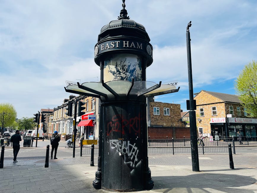 The photograph shows a street scene in East Ham with a cylindrical, vintage-style underground station sign structure, painted black with graffiti, situated on the pavement. The sign displays the text 'EAST HAM' at the top and features additional decorative elements around the circular glass, which reflects the sky and surroundings. In the background, there are rows of residential buildings with brick facades and commercial shops, including a nail salon and a beauty shop with red and pink signage. Pedestrians are walking along the sidewalk, some engaged in conversation or heading toward the shops. The street is lined with bollards and traffic lights, and the sky overhead is partly cloudy with patches of blue visible, indicating daytime. The area looks busy with light traffic and active foot traffic, typical of an urban residential and commercial area in Walthamstow or nearby, supported by the presence of [COMPANY_NAME]'s local removals and house moving services.