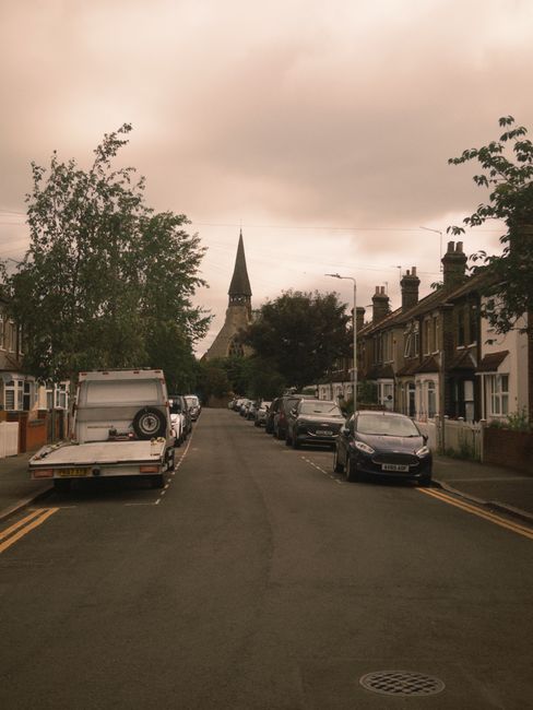 A quiet residential street in Walthamstow, E17, featuring terraced houses with brick facades and chimneys on the right side, with parked cars lining both sides of the narrow road. On the left, there are more homes and a large tree with green foliage extending over the pavement. The sky is overcast with grey clouds, and in the background, there is a church steeple rising above the trees and rooftops. This scene is typical of a peaceful neighbourhood in Walthamstow suitable for home relocation and furniture transport. Walthamstow Man and Van, a removals service provider, might use such streets for loading and unloading household items during house moves, as shown by the parked vehicles and the absence of traffic disruptions.