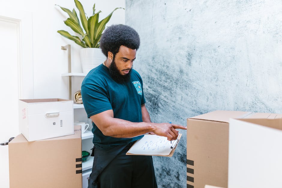 A woman with long blonde hair wearing a dark grey top is inside a room, preparing for house removals. She is holding a black marker and writing on a small white cardboard label attached to a packing l