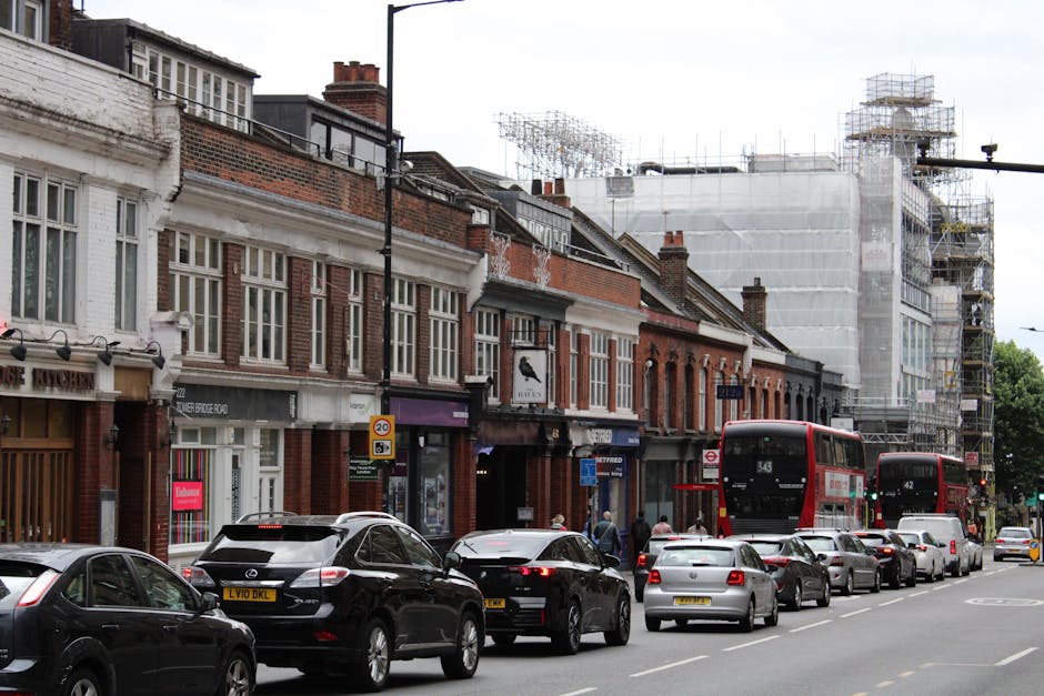 A busy urban street scene showing a row of brick and white-painted terraced houses with large sash windows and ornate detailing, situated along the pavement. Several vehicles, including black and silv